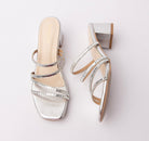 Silver criss cross strap mules for women, bright silver mirror-finish pu across the toe band and quarters, with four narrow straps sheathed in faceted silver stones that read almost like a row of tiny mirrors, a pale cream footbed with gold branding that contrasts against the cool silver upper, and a square block heel wrapped in matching silver metallic over a tan sole edge