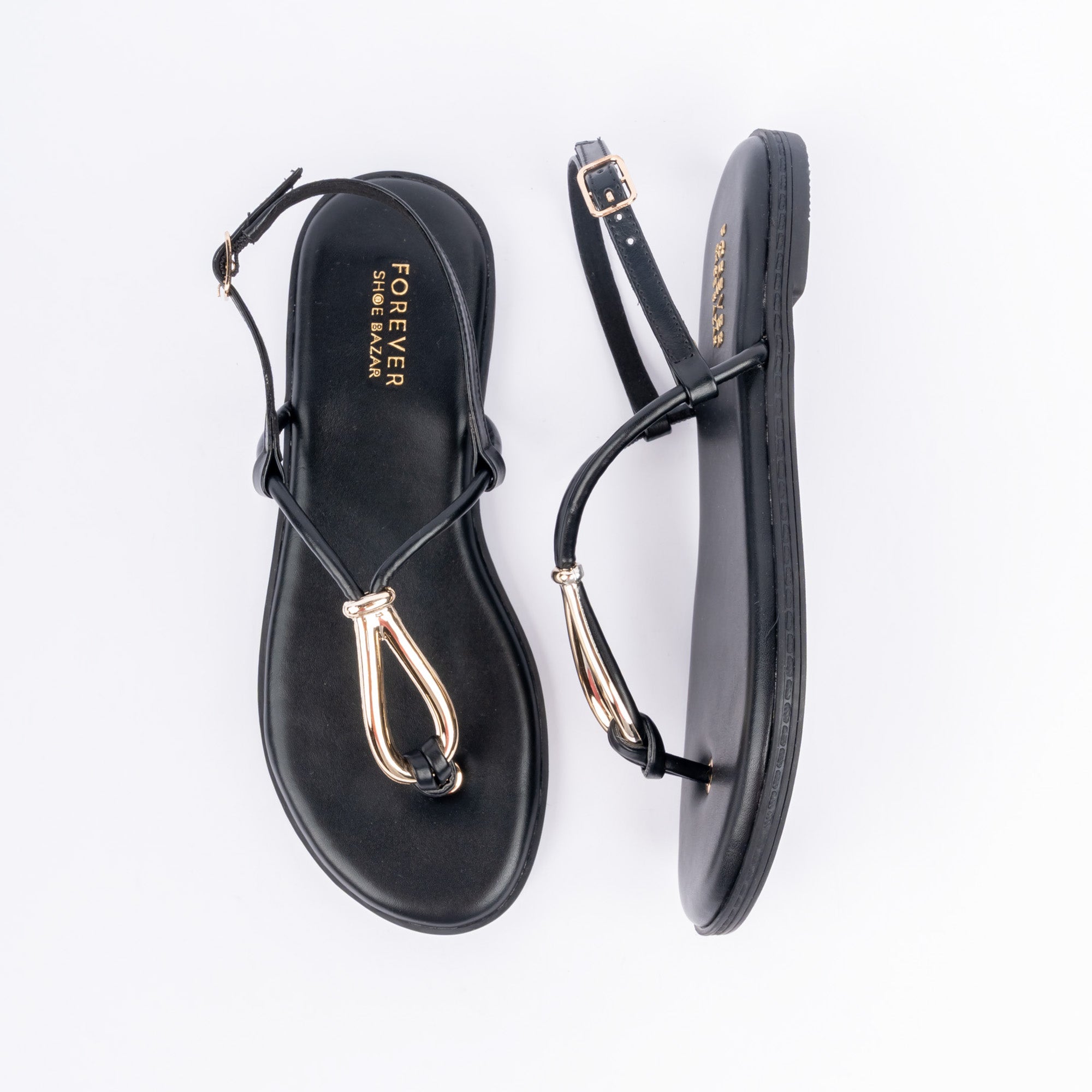 Black flat sandals for women, a slim toe-post capped with an elongated gold metal loop sitting flat on the vamp, feeding into a hairline strap round the heel to a gold pin buckle, on a cushioned footbed