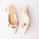 Beige rosette slingback pumps for women, an ivory pointed-toe slingback carrying a cluster of mauve and cream satin ribbon roses on green fabric leaves across the vamp, with an adjustable silver pin-buckle sling strap and a flared spool kitten heel above a padded tonal footbed