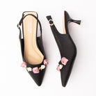 Black rosette slingback pumps for women, a black pointed-toe slingback trimmed with a row of dusty-pink and ivory satin ribbon roses on green fabric leaves across the vamp, with a silver pin-buckle sling strap and a flared spool kitten heel over a contrast cream cushioned footbed