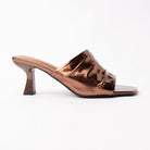 Bronze metallic mules for women, one wide panel of mirror-foiled bronze leather gathered across the instep and caught underneath by a slim piped strap over the toes, open through the arch and heel, on a slim flared spool heel in matching antique-copper foil