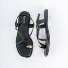 Black flat sandals for women, a polished gold nugget capping the big-toe loop, two slim straps crossing the vamp, and a buckled backstrap over a cushioned footbed on a low tread sole