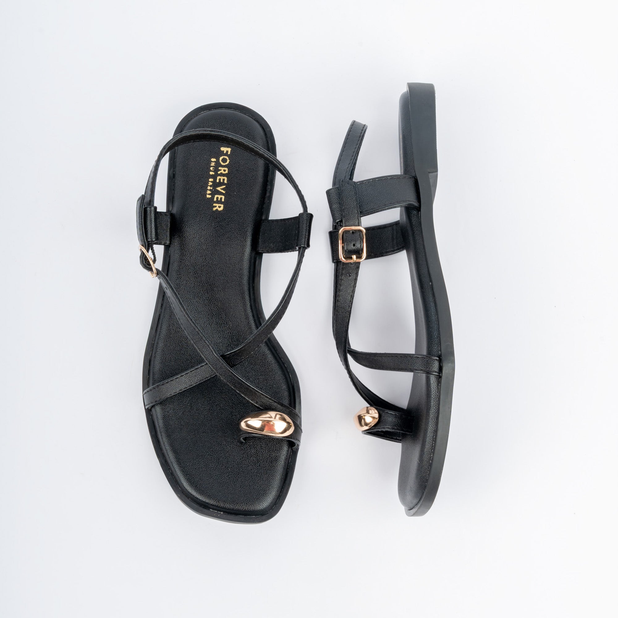 Black flat sandals for women, a polished gold nugget capping the big-toe loop, two slim straps crossing the vamp, and a buckled backstrap over a cushioned footbed on a low tread sole