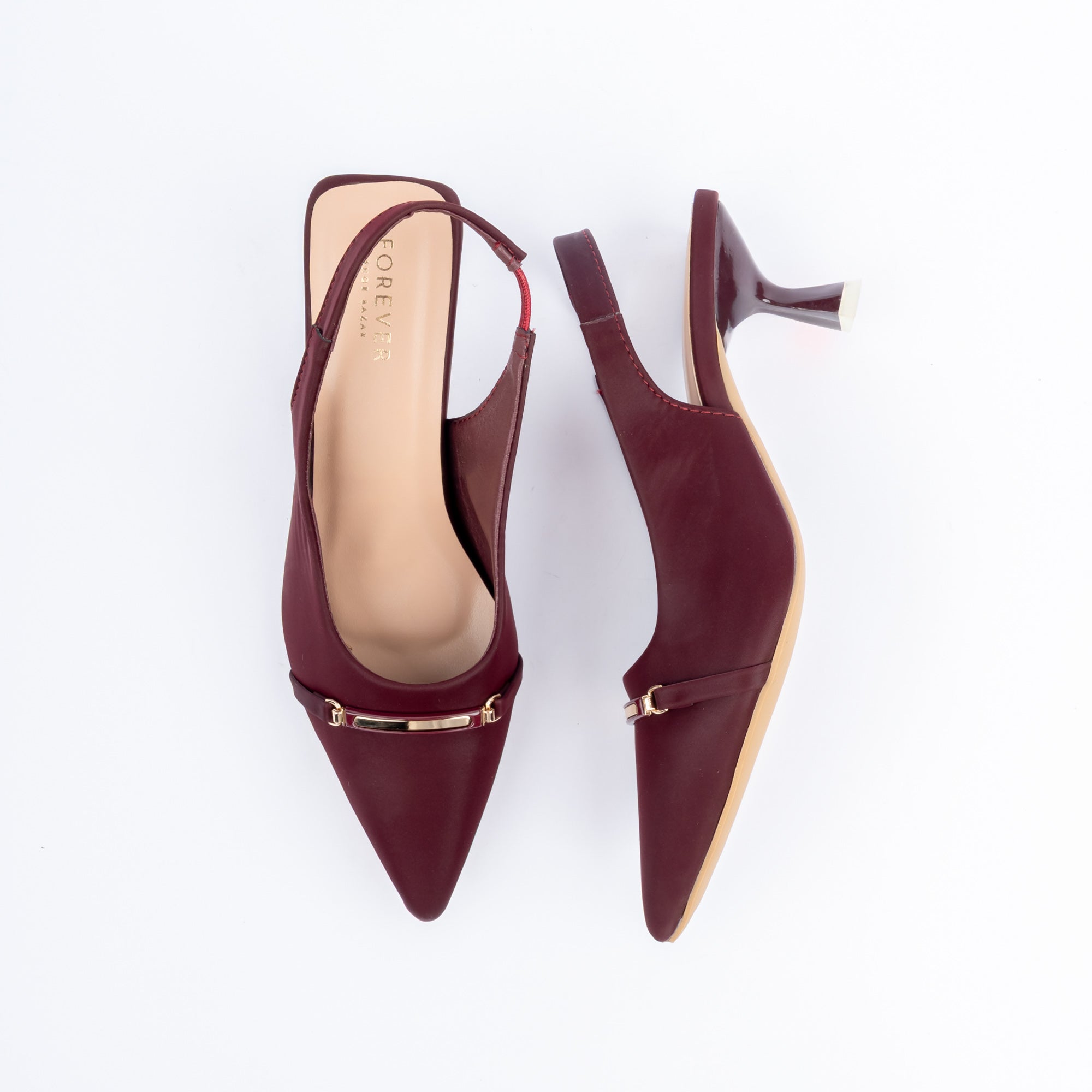 Maroon pumps for women, a slim gold bar plaque anchored by a small buckle fitting on a narrow strap across the pointed vamp, with an elastic gusset set into the heel strap and a flared spool heel